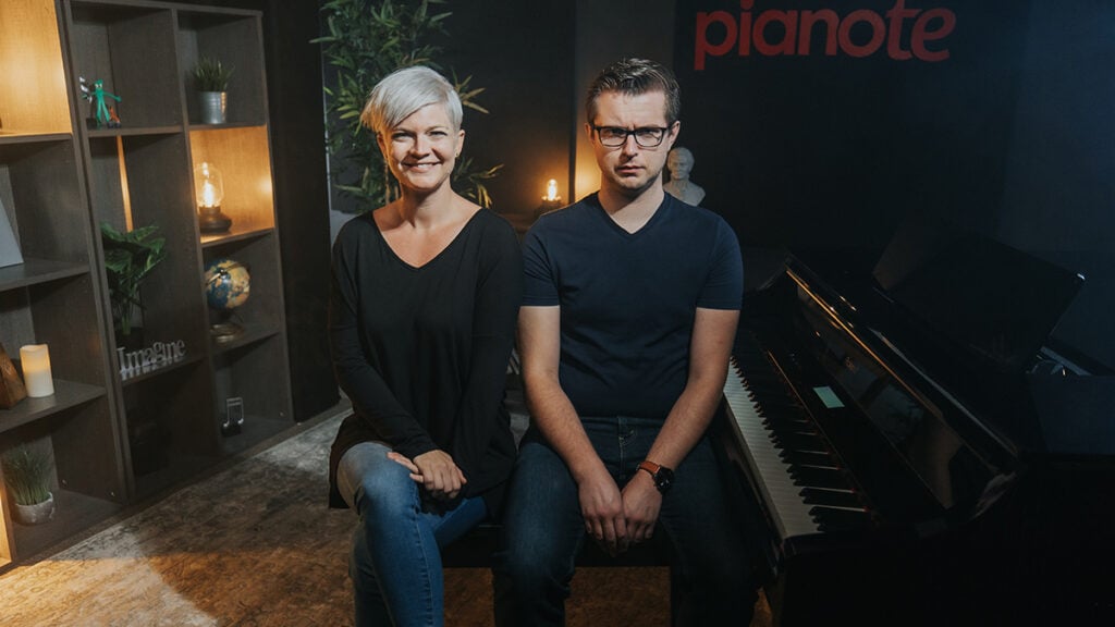 Sad and happy chord progressions. Smiling woman with short platinum hair smiling next to frowning man with glasses and brown hair on piano bench next to a grand piano.