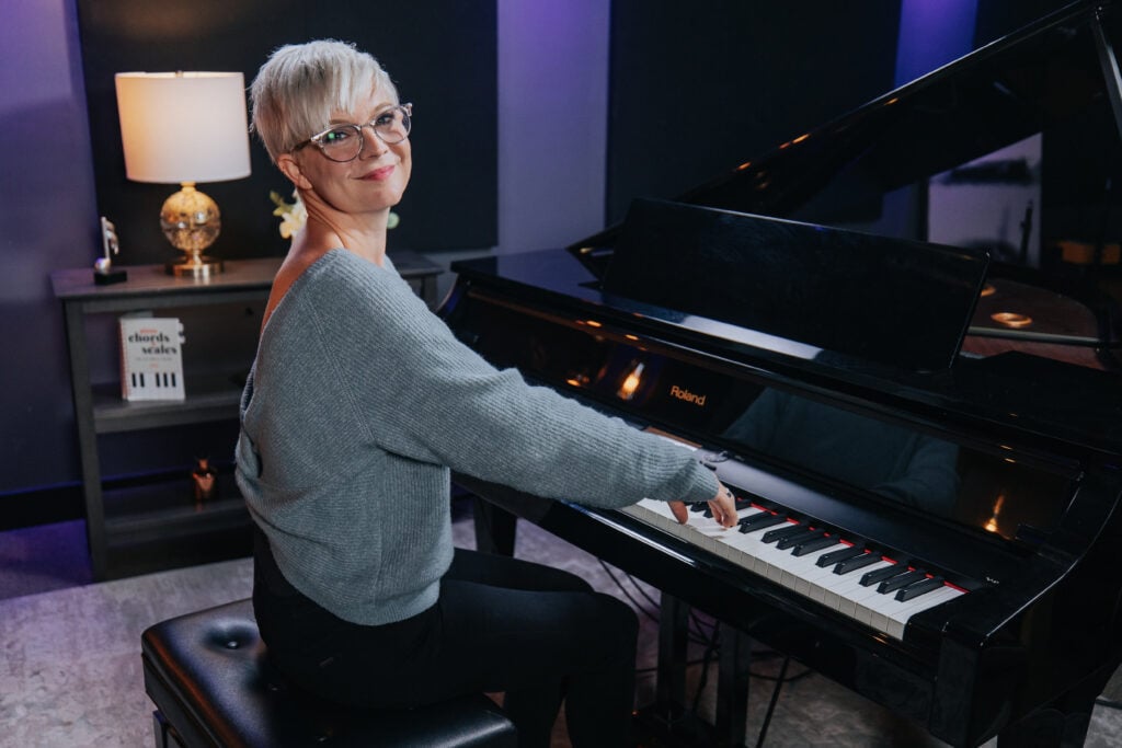 Woman with short platinum hair and light blue sweater playing grand piano in dark blue studio.