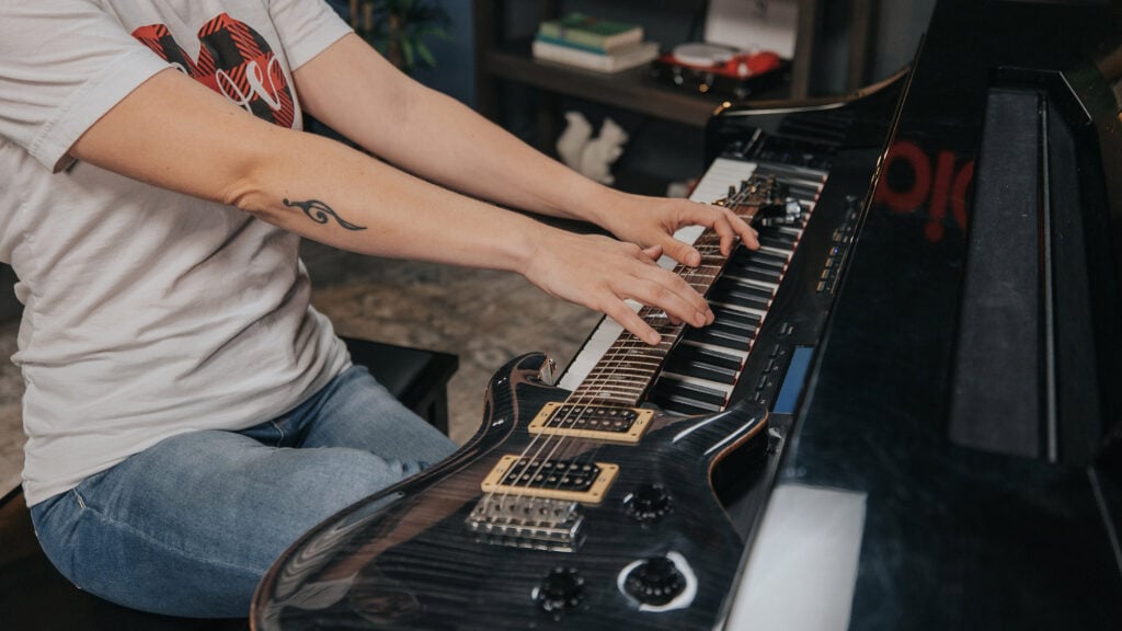 Electric guitar laid on a piano keyboard with hands playing on top.