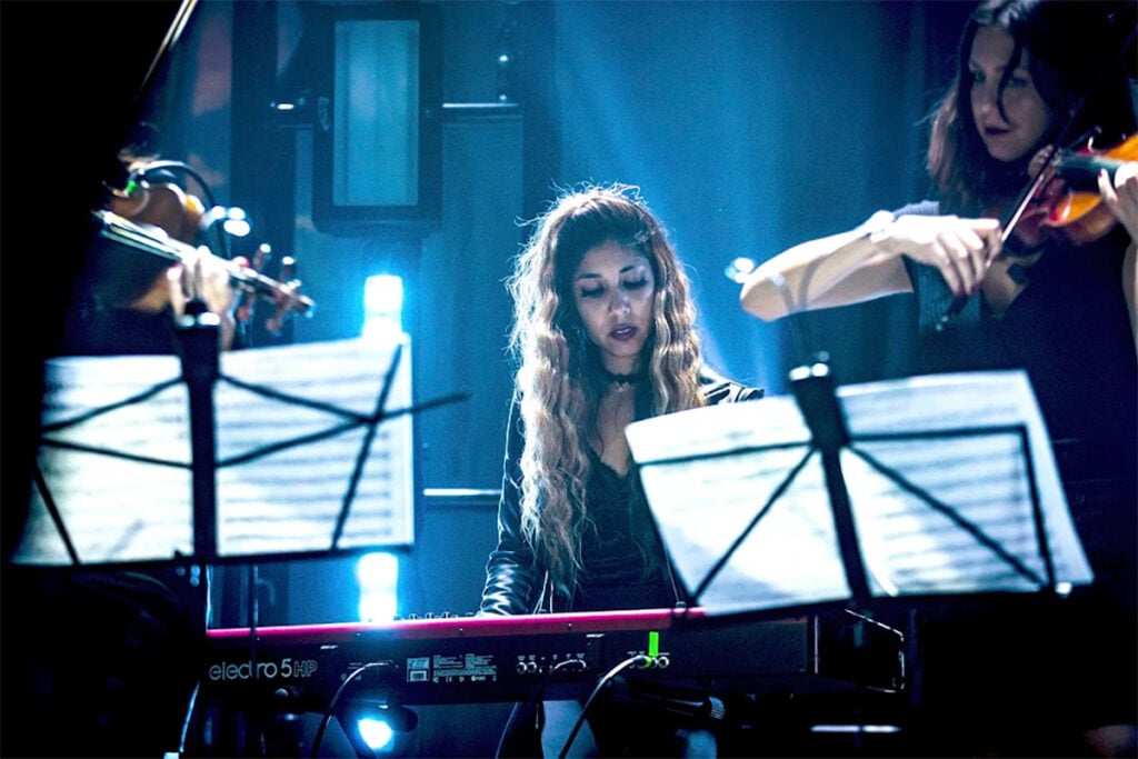 Woman with long curly hair sitting at red keyboard on stage next to violinists in blue neon lighting.