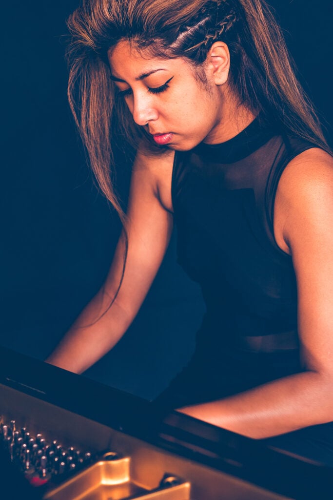 Summer Swee-Singh - young woman with amber-highlighted hair in black dress behind grand piano, playing, with inside of piano slightly exposed.