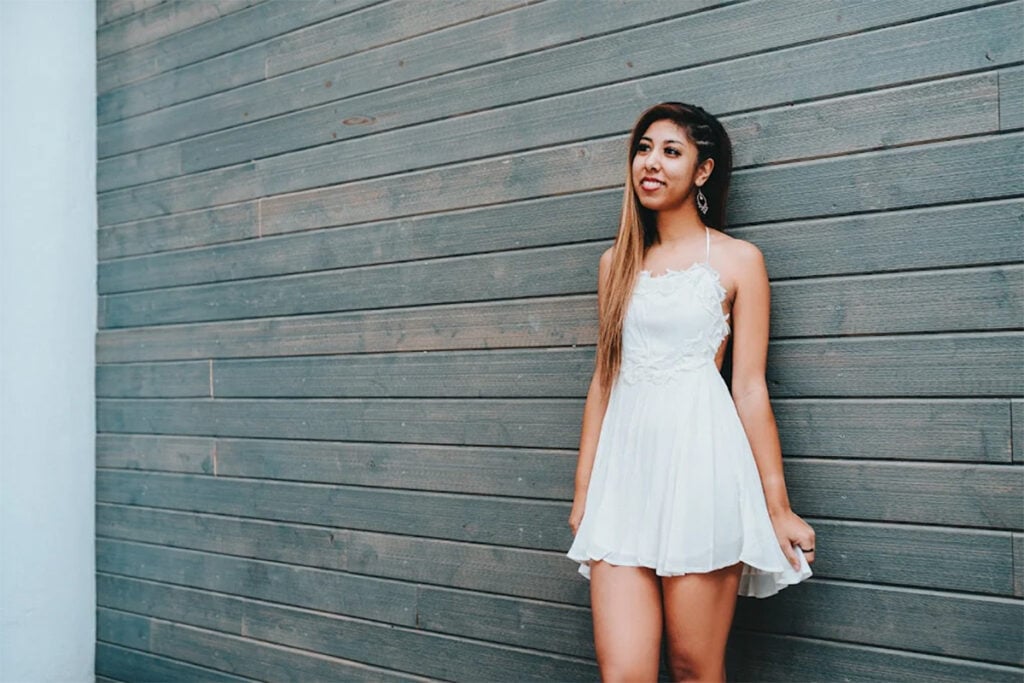 Summer Swee-Singh - young woman with long amber highlighted hair in short white dress smiling and standing against wooden plank wall.