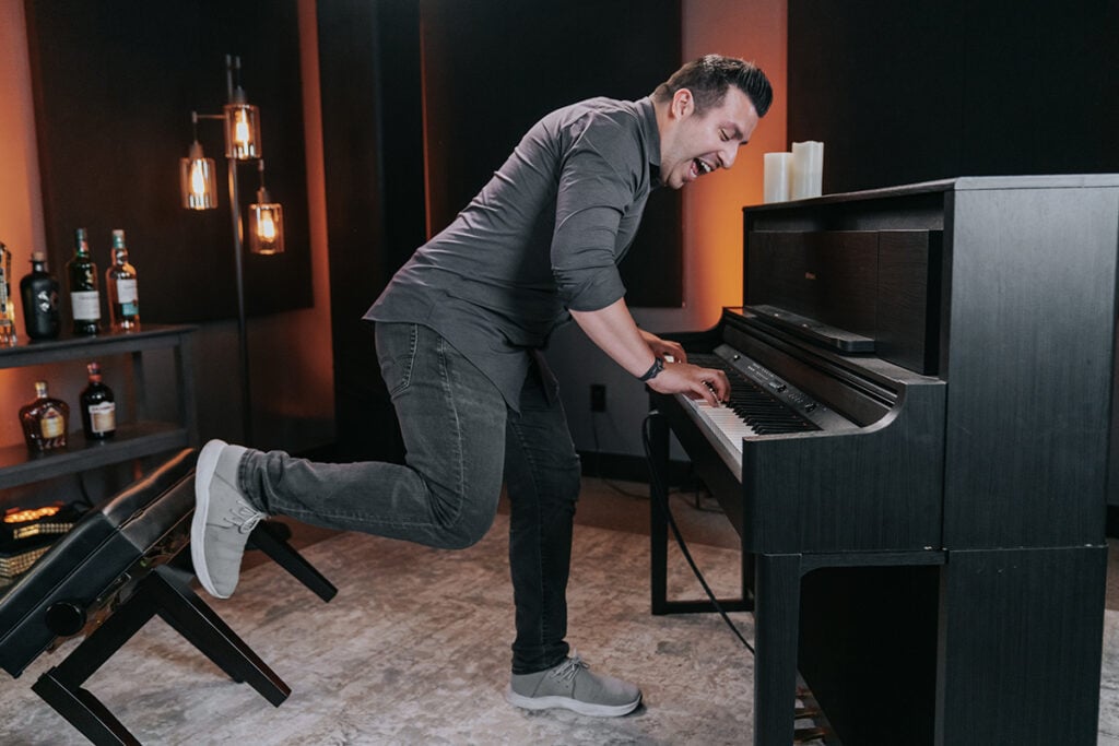 Man in grey shirt playing upright piano standing up and kicking back piano bench, with a bar in the background.