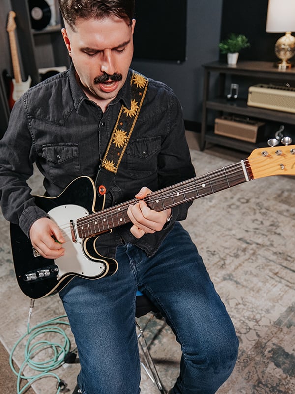 Man in black shirt and moustache playing black electric guitar, bending string.