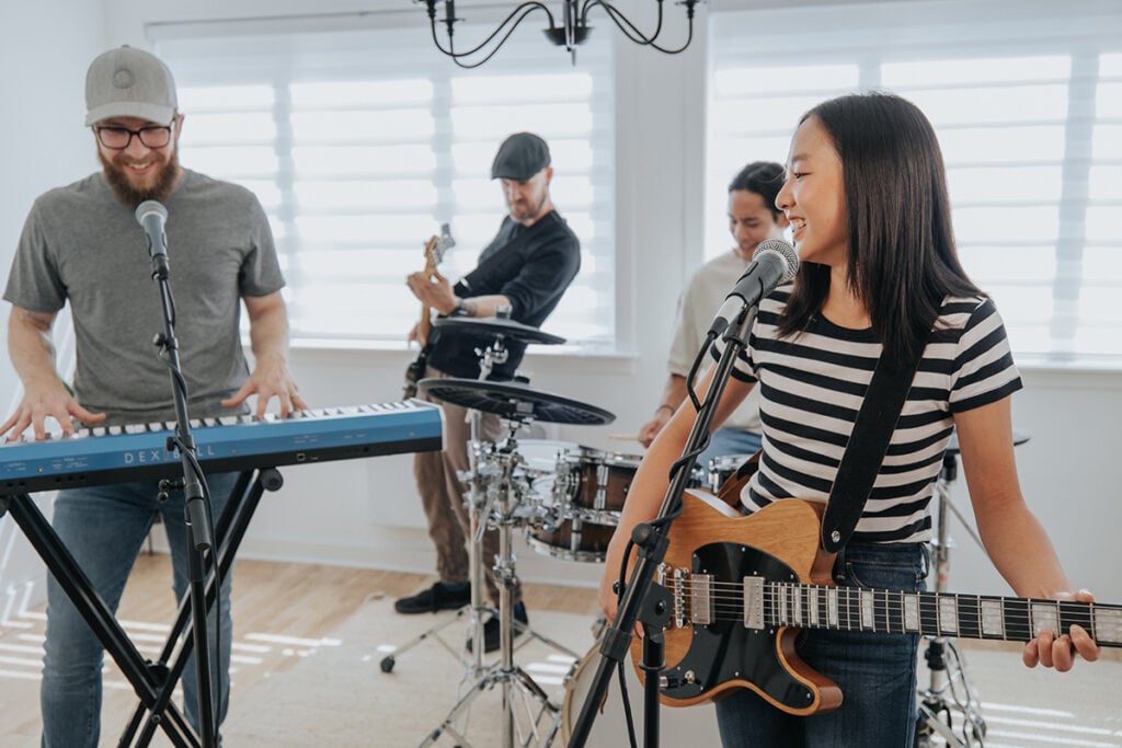 Band: man with beard and cap playing blue keyboard, young woman in stripey shirt playing electric guitar. Background: man in black cap playing bass, man in white shirt playing drums.