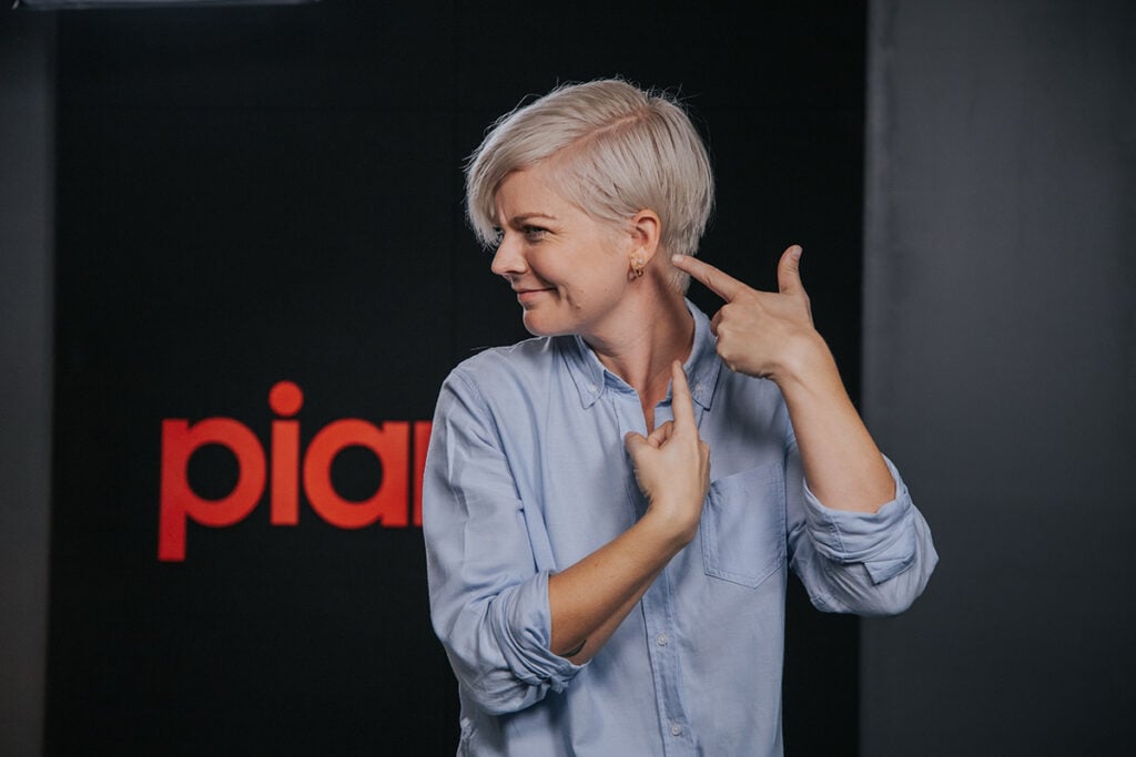 Woman with short platinum hair and light blue button down shirt pointing at ear and looking to the side.