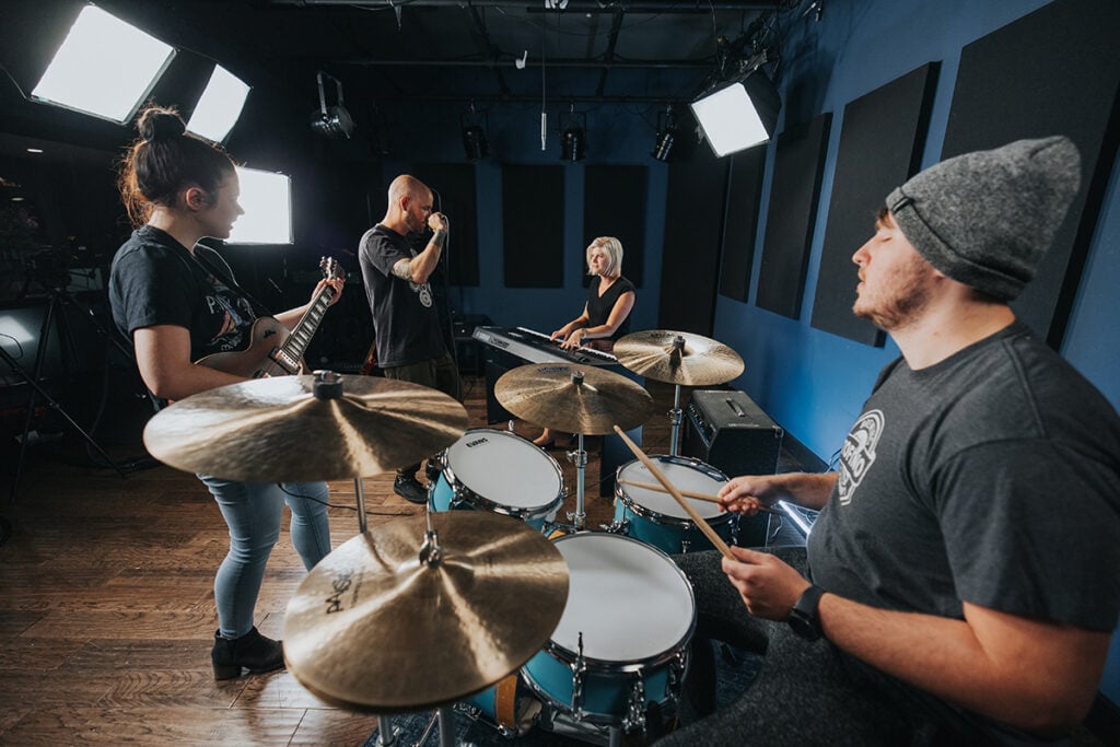 Drummer, keyboardist, vocalist, and guitarist playing in a blue studio.