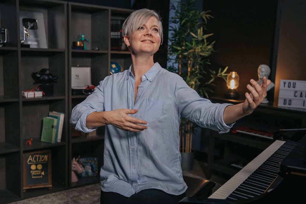 Woman with short platinum hair and light blue button down shirt using arms to express musicality in front of a piano.