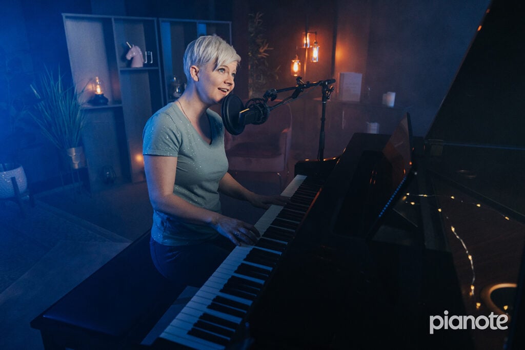 Woman with short platinum hair singing and playing piano in a dark blue lit studio