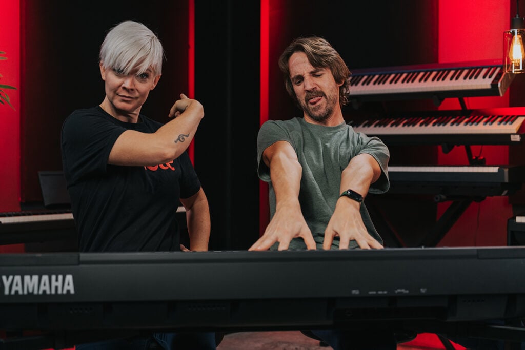 Woman with short platinum hair in black tshirt pointing behind at rows of keyboards and man with green shirt playing keyboard beside her with rock out expression.