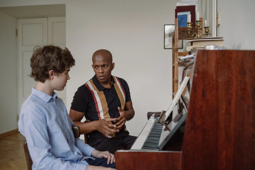 Man in stripey shirt sitting next to and talking to teen boy in blue button down in front of a piano.