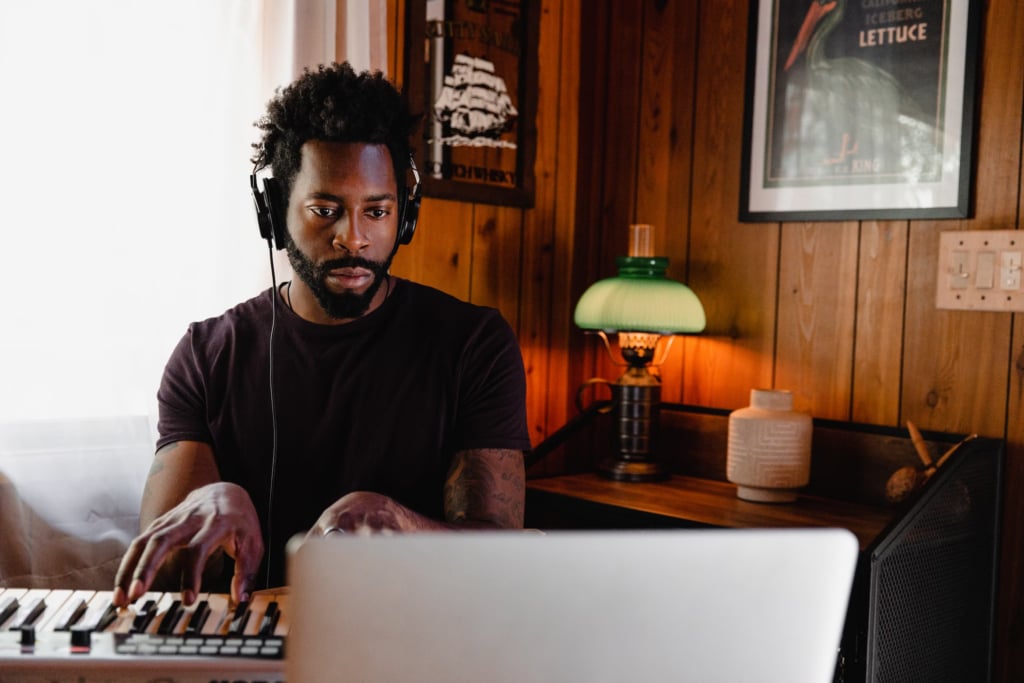 Man with dark beard playing keyboard with headphones in front of a laptop.