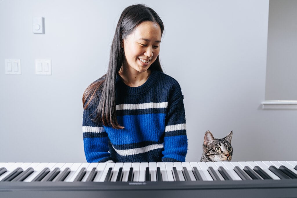 Is piano hard to learn? Woman with long dark hair in blue stripey sweater sitting behind piano keyboard with grey tabby cat next to her, head peeking out from behind the keyboard.