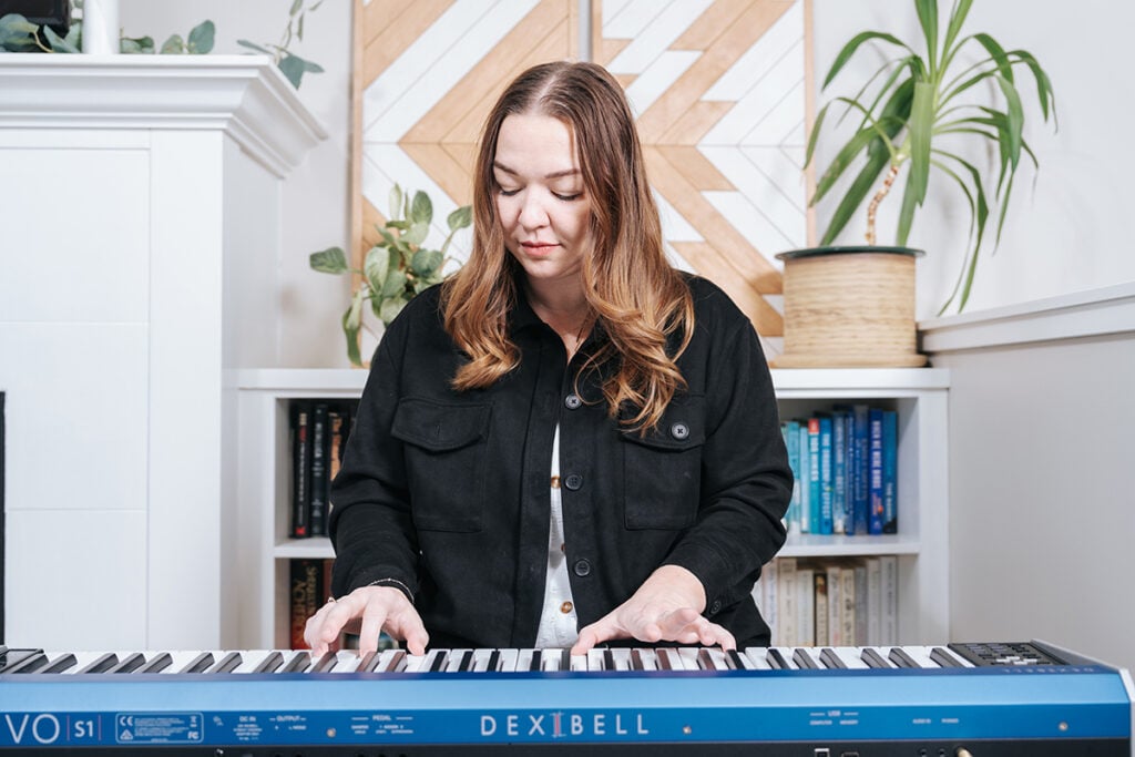 Is piano hard to learn? Woman with dark blonde hair playing blue keyboard in front of bookshelf with plants.