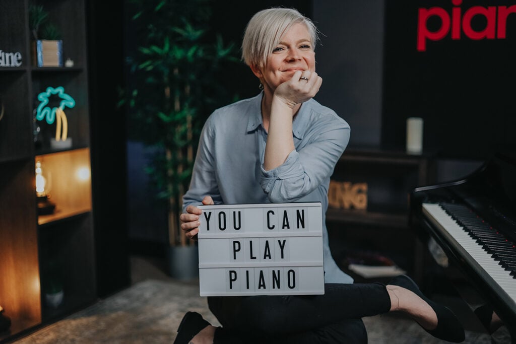 Woman with short platinum hair and blue blouse sitting next to grand piano cross legged with sign that reads YOU CAN PLAY PIANO.