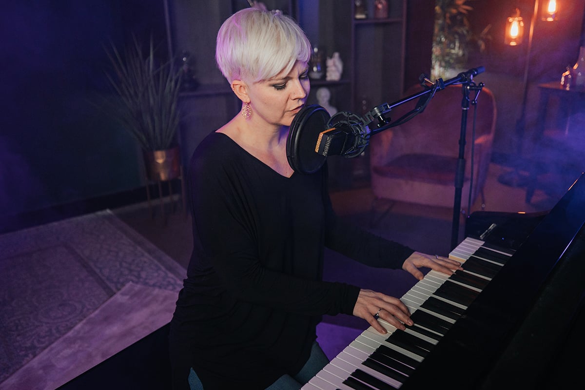 How to write a song on piano. Woman with short platinum hair playing piano and singing in mic in studio with purple lighting.