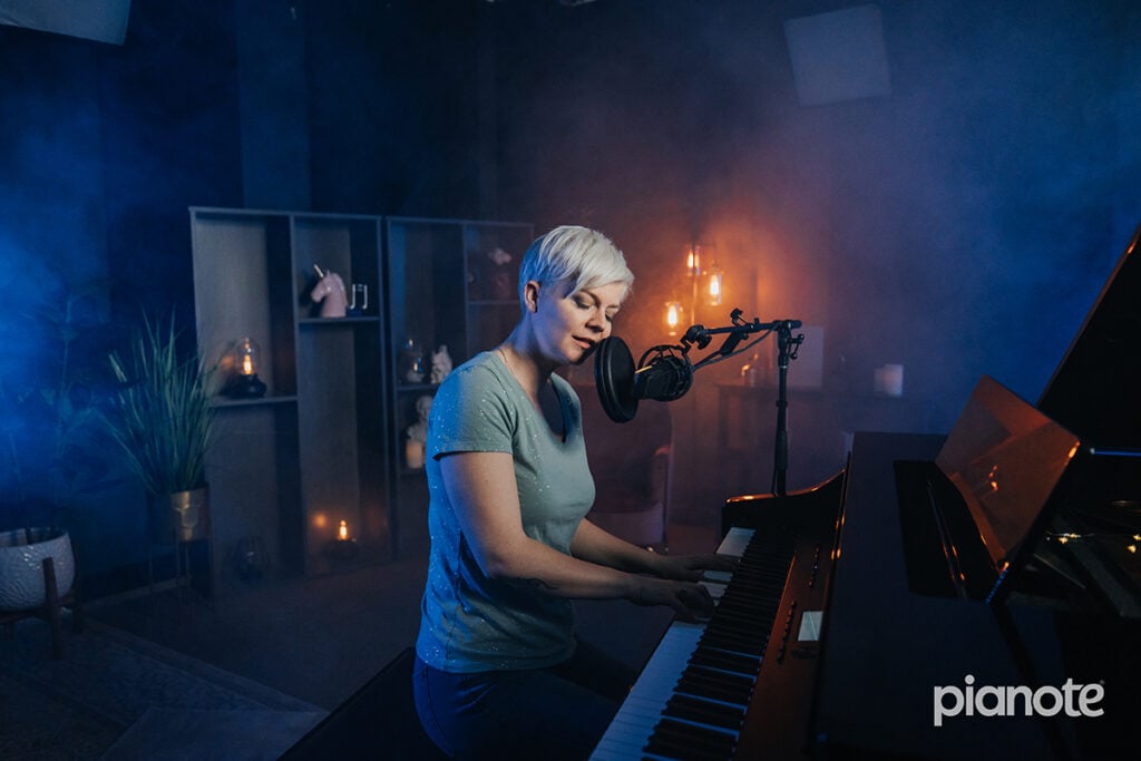 Woman with short platinum hair playing grand piano in blue lit studio with sparkly blue shirt singing into microphone.