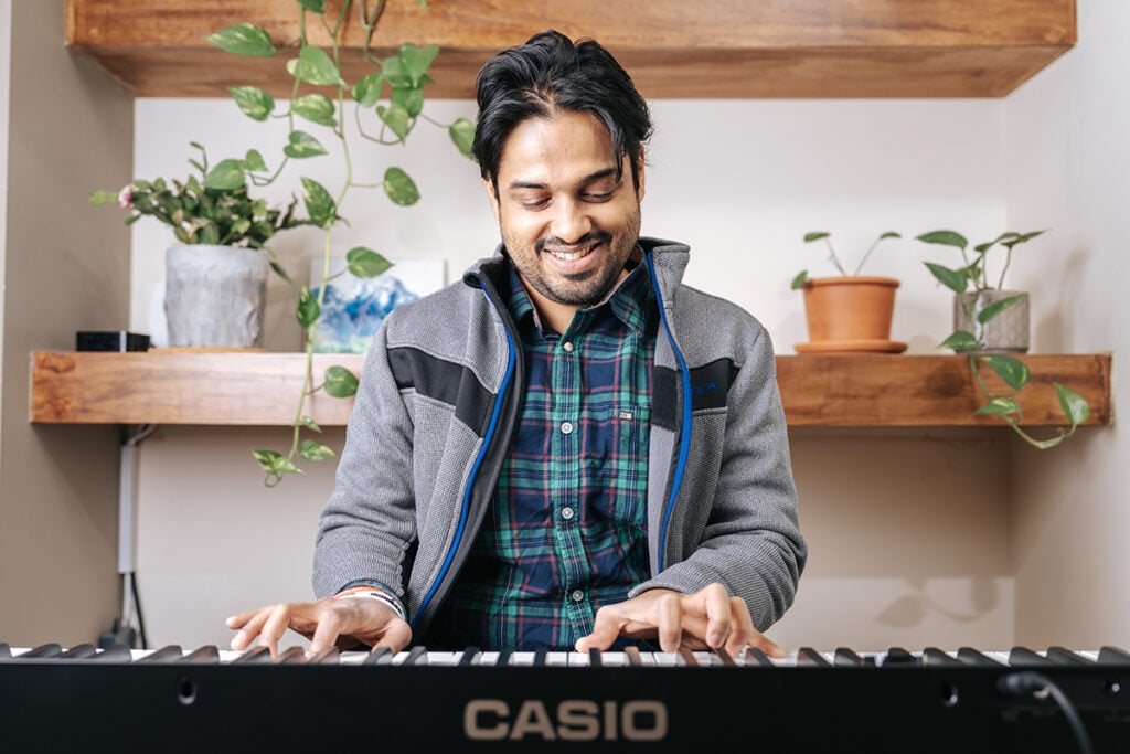Man in green blue plaid shirt and grey sweater playing casio keyboard.