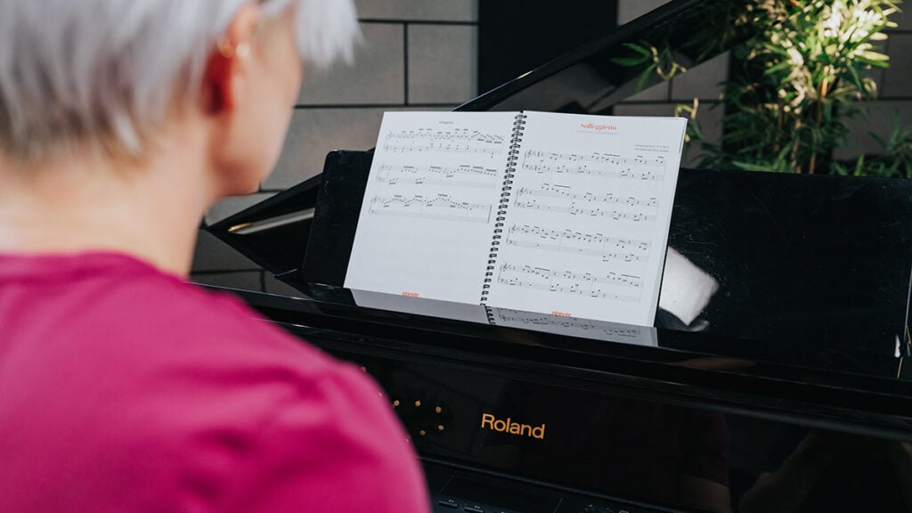 Behind shoulder shot of woman with short platinum hair reading sheet music on piano music stand.