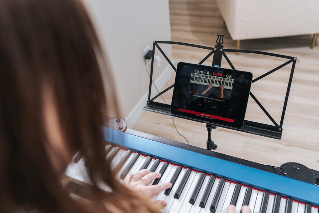 Over the shoulder shot of person with long hair playing blue keyboard with iPad showing online lesson on music stand.