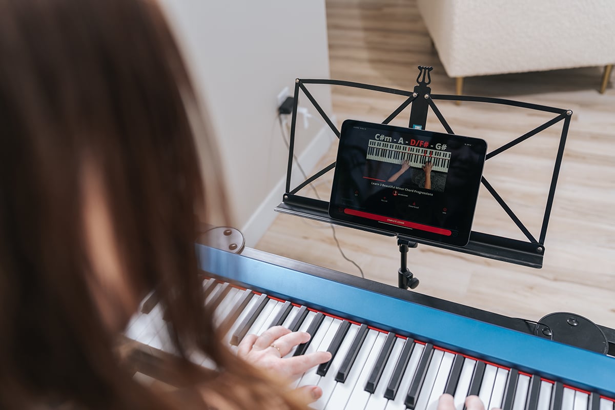 Over shoulder shot of person with long hair playing blue keyboard with iPad showing piano lesson on music stand.