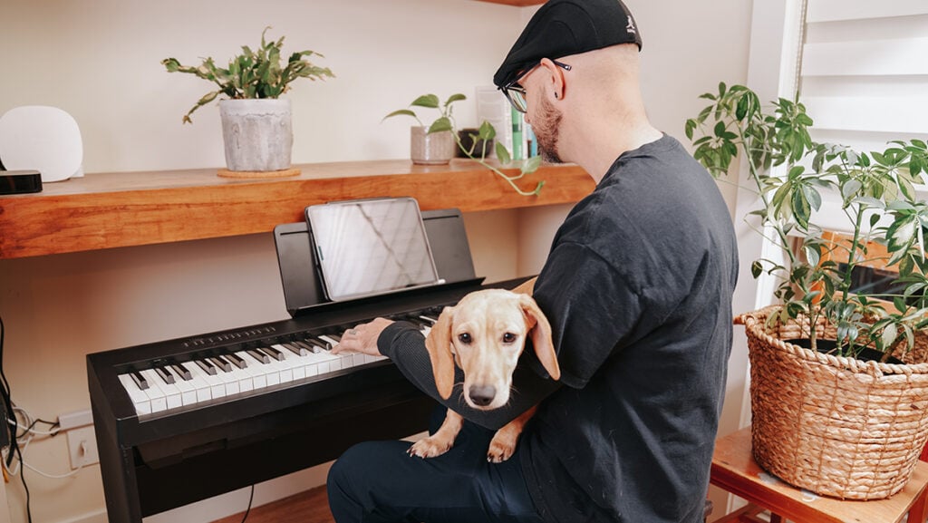 Man with hat playing keyboard with iPad on piano stand with dog in lap.