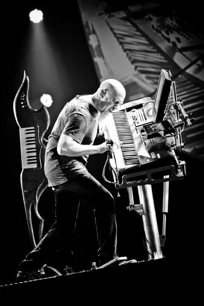 Black and white photo of man with goatee playing keyboard standing up on stage.