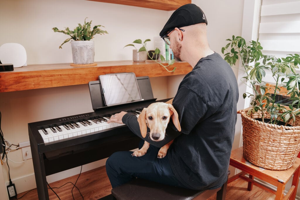 Man with hat playing keyboard with sausage dog on lap.