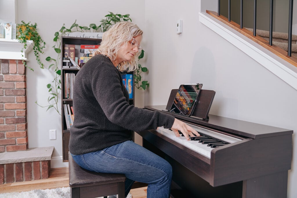 How long should you practice piano? Woman with medium blonde hair playing upright keyboard piano against stairs in home.