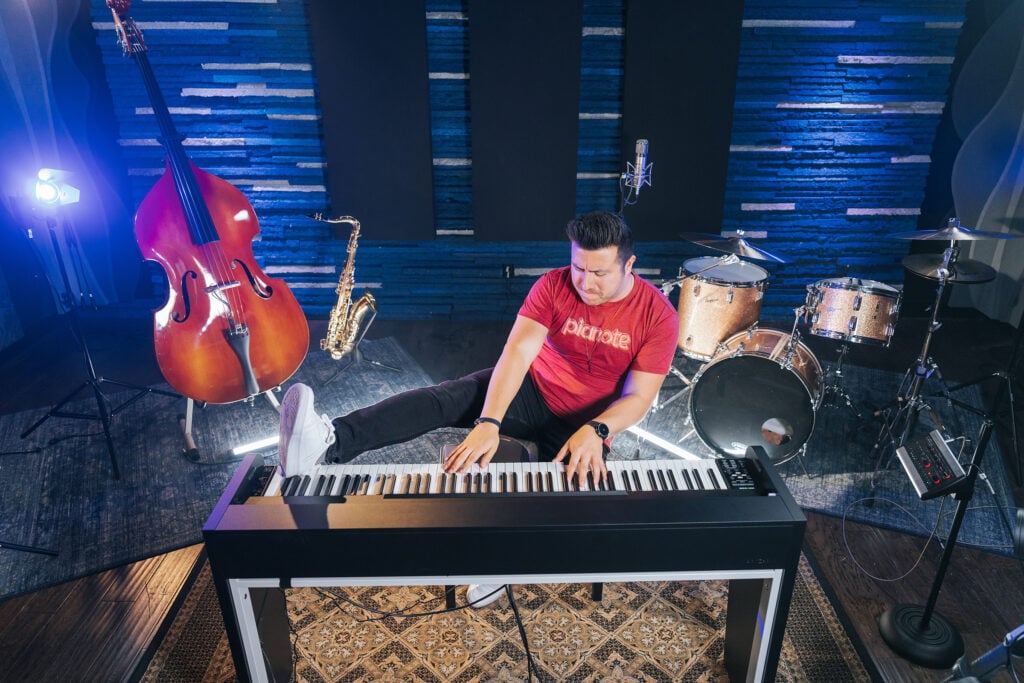 Man in red shirt playing keyboard standing up with foot on keys in front of drums, microphones, double bass, and saxophone.