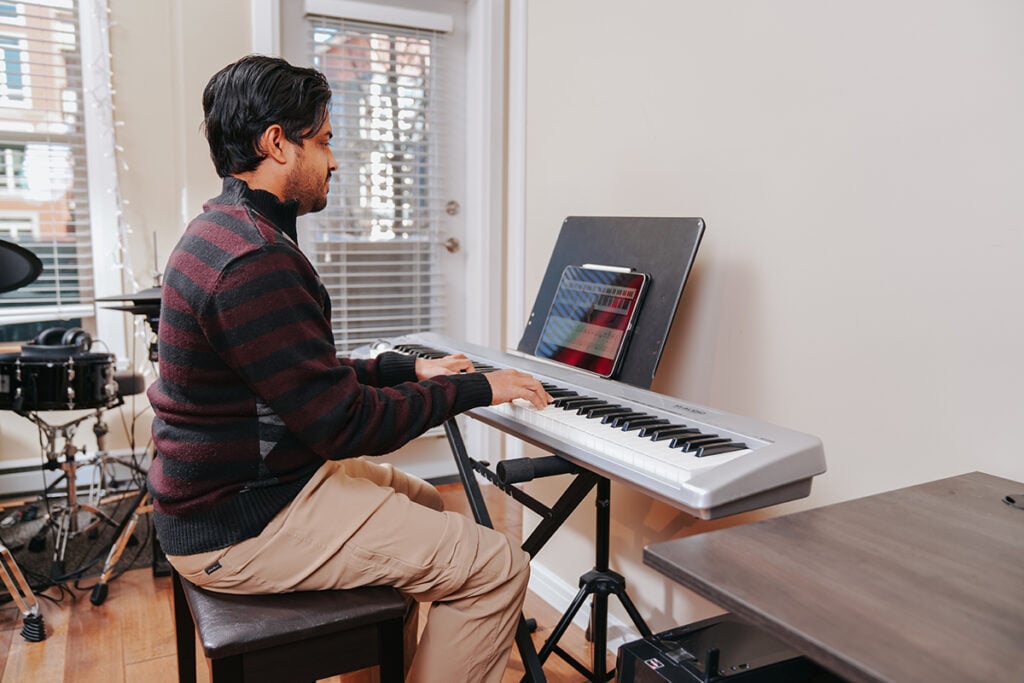 Man with dark hair in khaki pants and stripy sweater playing silver keyboard with iPad on music stand and drum set in the background.