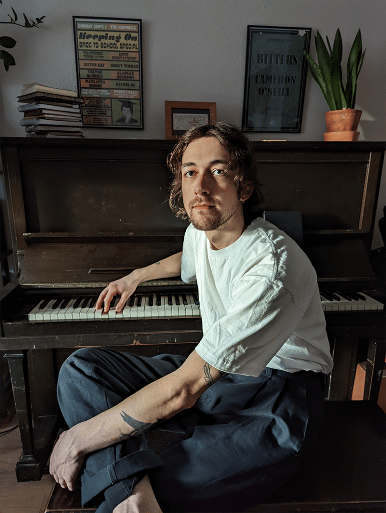 Man in white shirt sitting in front of old upright with one hand on keys.