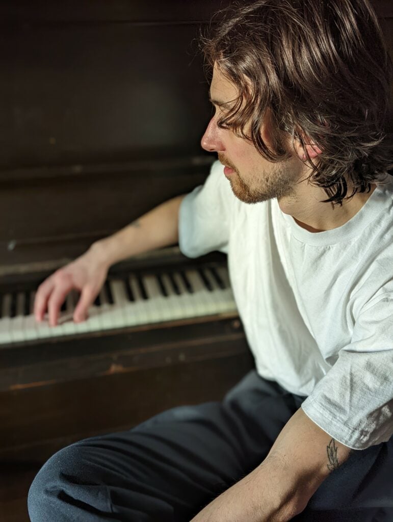 Man with medium length hair in white shirt looking at upright piano with one hand on keys.