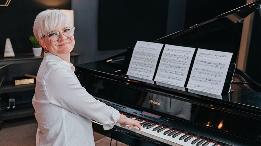 Woman with short platinum hair playing grand piano looking at camera over shoulder, with sheet music on piano stand.