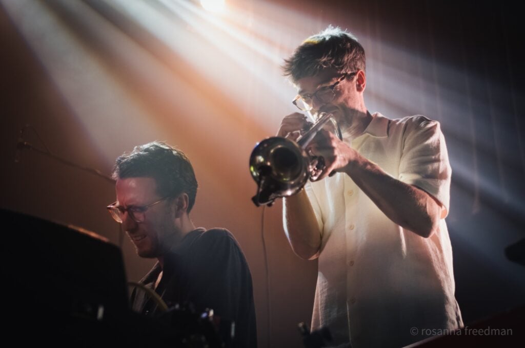 Man with glasses in white button shirt playing trumpet on stage behind another man with glasses playing keyboard.