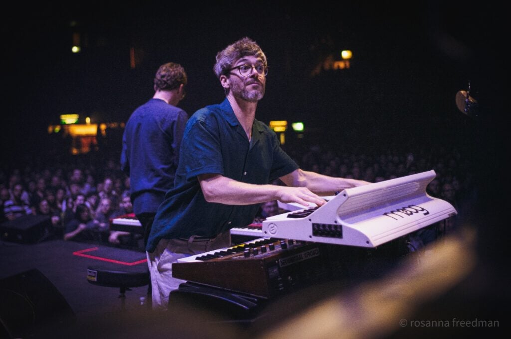 Man with glasses playing white keyboard on stack of keyboards on stage.