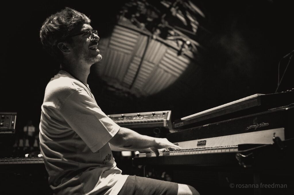 Black and white photo of man in glasses playing Rhodes piano next to other synthesizers.