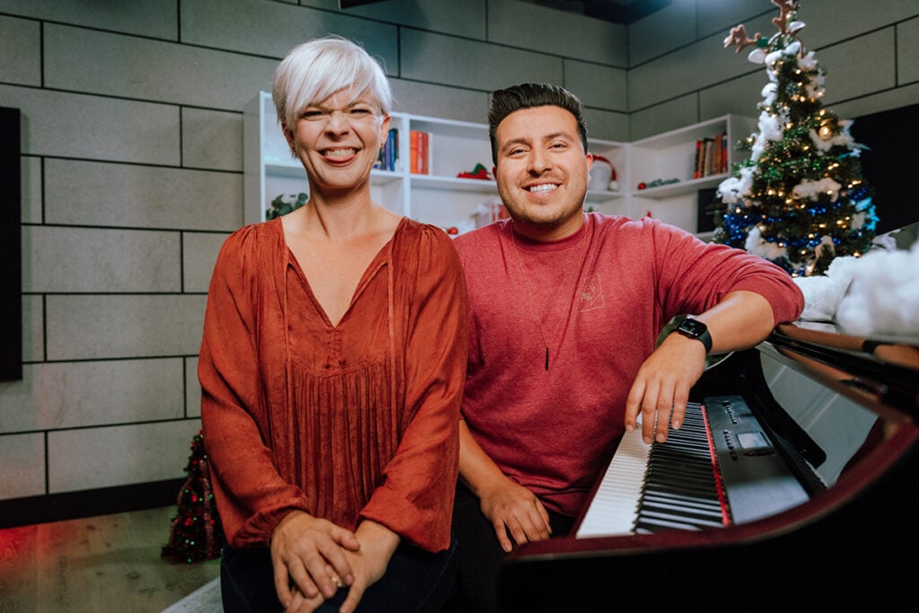 Woman with short platinum hair and man with short dark hair both in red sitting on piano bench next to piano in front of Christmas tree in studio. Woman makes silly face.