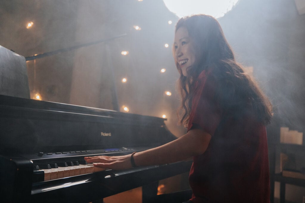 Woman with long hair and red shirt playing grand piano in smoky studio.