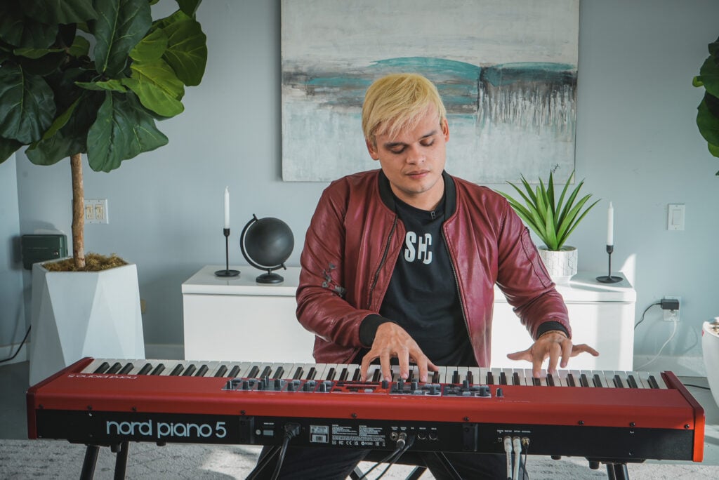 Man in red jacket and dyed blond hair playing red keyboard in swanky modern apartment.