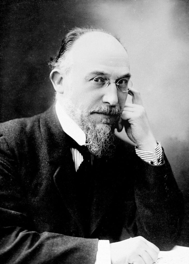 Monotone photo of man with scraggly beard and small glasses in suit.
