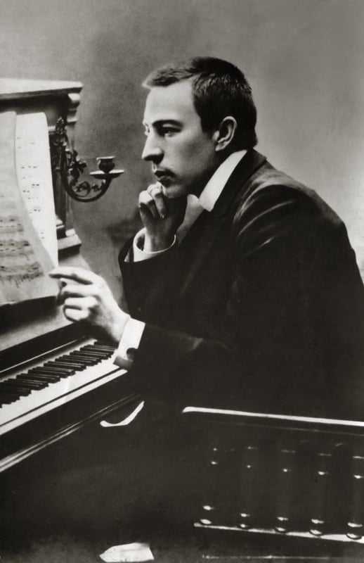 Monotone photo of man with short hair in suit looking at sheet music at an upright piano.