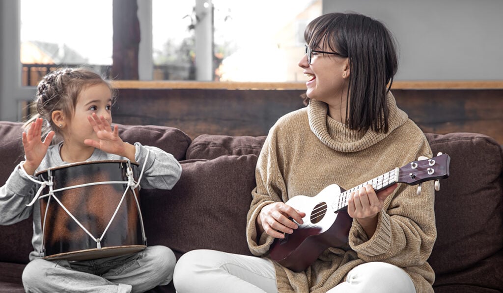Girl and woman on brown couch. Girl plays drum with hands, woman sings and plays ukulele.