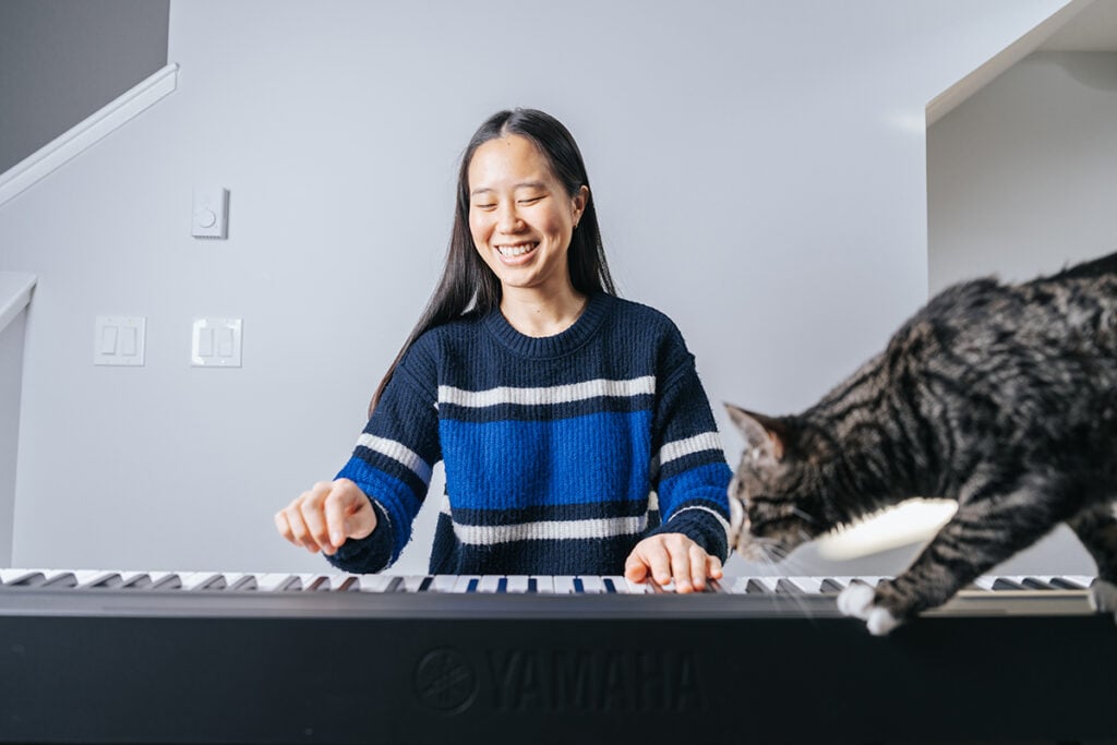 Woman with long hair and blue sweater playing digital piano while tabby cat walks across.