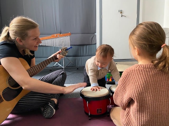 Woman with pin stripe pants holding guitar sitting next to toddler playing drums and young girl.