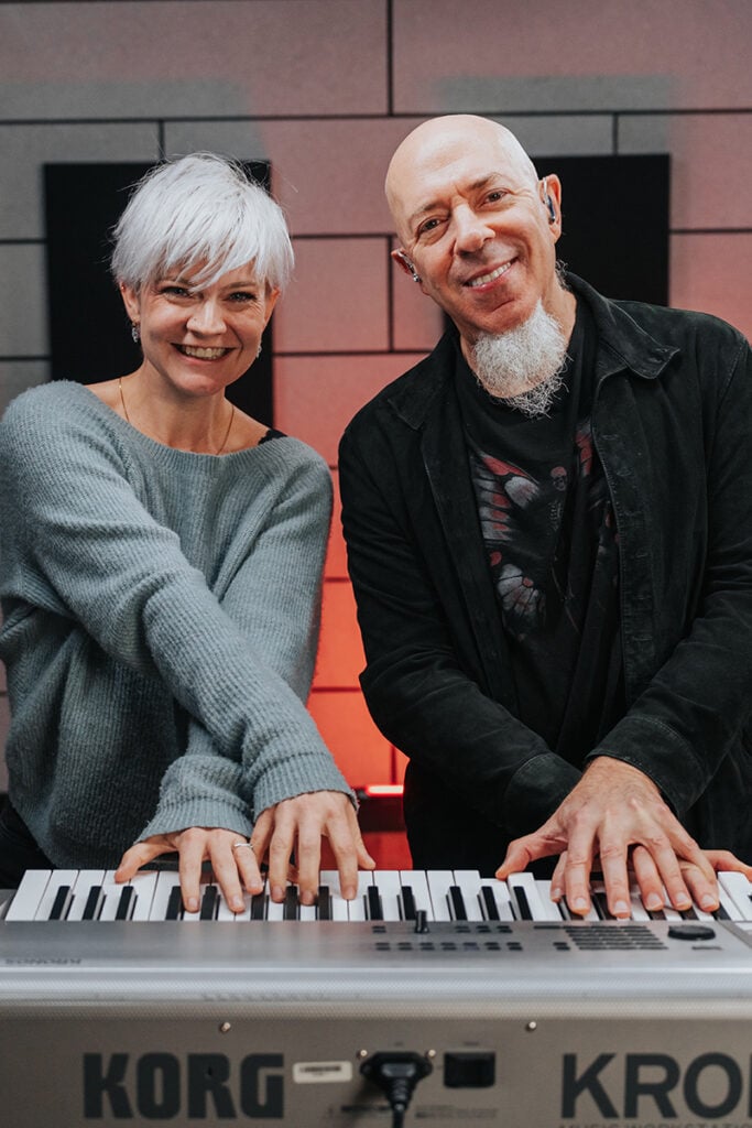 Woman with short platinum hair and man with white goatee at a piano with their hands crossed.