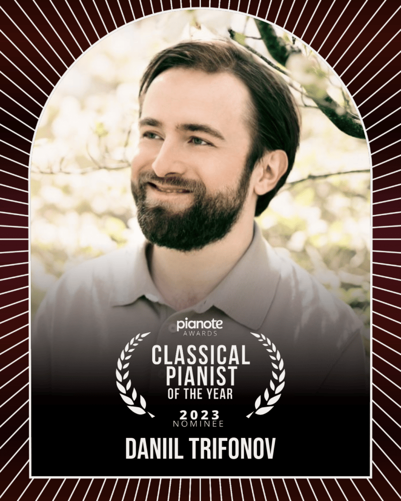 Daniel Trifonov. Smiling man with beard in collared shirt in front of light coloured trees.