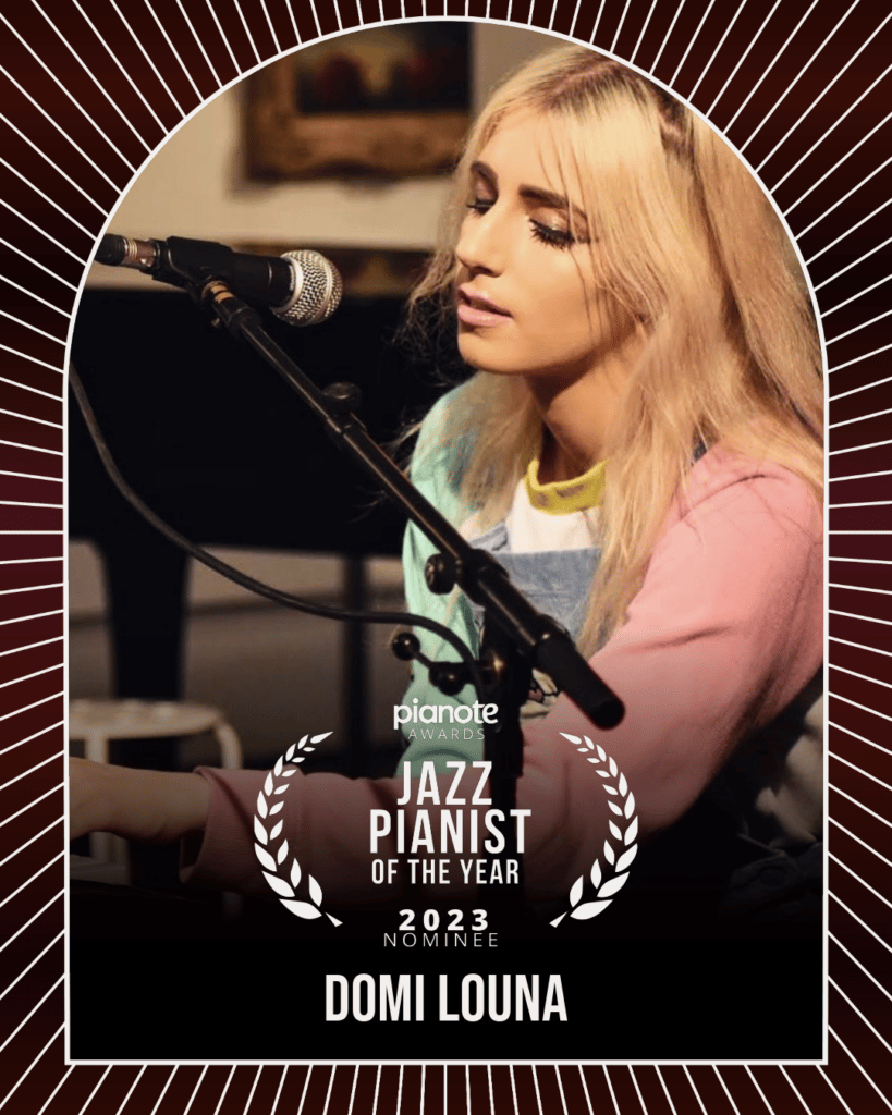 DOMi Louna. Young woman with long blonde hair and pastel clothing playing keyboard in front of a mic.