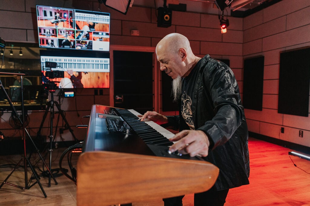 Man with goatee playing keyboard in studio with screens.