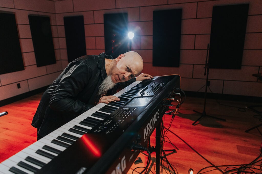 Man with goatee in leather jacket playing and leaning over keyboard in studio.